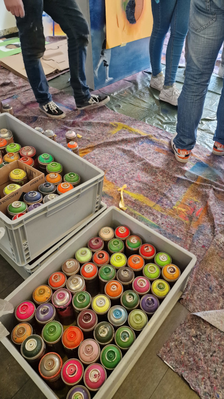 Zwei Kisten voller Farbflaschen die auf dem Boden stehen. Im Anschnitt sieht man die Beine von drei Menschen.
